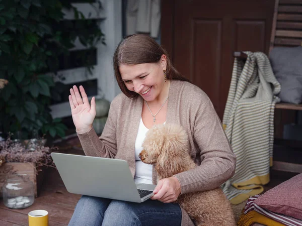 middle aged caucasian woman in brown cardigan sits on terrace of house with her beige poodle with laptop working outdoors in garden, home office , Education, modern lifestyle and leisure concept.