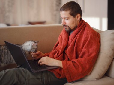 Ill man covered in warm blanket looking at laptop screen, sitting on sofa at home, empty space. Sinusitis illness, cold and influenza symptoms. Working remotely at home.