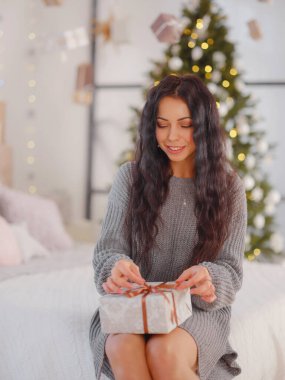 Yatak odasındaki noel ağacının yanında hediye kutusuyla gülümseyen genç kadın. Tatil kavramı. Gri kazaklı beyaz bir kadın. Noel Ağacı süsleme. Noel sabahında hediyeleri açmak