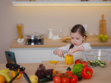 Genç Vlogger kız evde yemek pişirme hakkında sosyal medya videosu yapıyor, tripodla akıllı telefona bakıyor. Sağlıklı beslenmeyle ilgili bölüm. Çocukları erken yaşta sağlıklı beslenmeye çekme kavramı