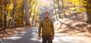 Genç yakışıklı adam sonbahar ormanında poz veriyor. Sırt çantalı genç hippi, ormanda duran gezgin, yürüyüş, orman, yolculuk, aktif sağlıklı yaşam tarzı, macera, tatil konsepti..