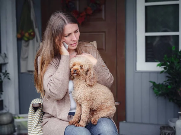 middle aged caucasian woman in brown cardigan sits on terrace of house with her beige poodle and emotionally speaks with friend via smartphone. morning good and bad news, autumn lifestyle concept