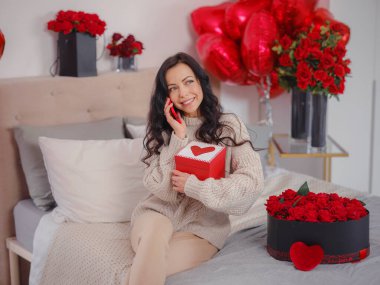 Gülümseyen mutlu kadın, hediye kutusuyla telefonda sevdikleriyle konuşuyor, yatakta oturup kameraya bakıyor. Sevgililer Günü 'nün kutlaması, düğün ve aşk günü, kadınlar günü. Sabah aşk yatak odası.