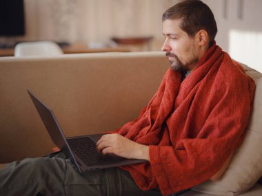 Sıcak battaniyeye sarılı bir adam dizüstü bilgisayara bakıyor, evdeki kanepede oturuyor, boş bir alan. Sinüzit hastalığı, soğuk algınlığı ve grip belirtileri. Evde uzaktan çalışıyor..