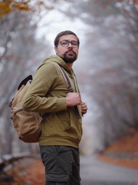 Genç yakışıklı adam sonbahar ormanlarında yolda poz veriyor. Sırt çantalı genç hippi, ormanda duran gezgin, yürüyüş, orman, yolculuk, aktif sağlıklı yaşam tarzı, macera, tatil konsepti..