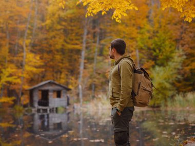 Genç yakışıklı adam göl kenarındaki sonbahar ormanında poz veriyor. Sırt çantalı genç hippi, ormanda duran gezgin, yürüyüş, orman, yolculuk, aktif sağlıklı yaşam tarzı, macera, tatil konsepti..