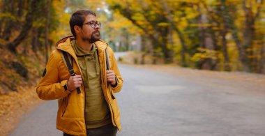 Genç yakışıklı adam sonbahar ormanında poz veriyor. Sırt çantalı genç hippi, ormanda duran gezgin, yürüyüş, orman, yolculuk, aktif sağlıklı yaşam tarzı, macera, tatil konsepti..