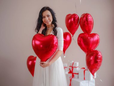 Beyaz elbiseli güzel, mutlu genç bir kadın. Tatil partisi. Kırmızı kalp şeklinde balonlarla poz veren neşeli manken, eğlenen, Sevgililer Günü 'nü kutlayan. Aşkın sembolü