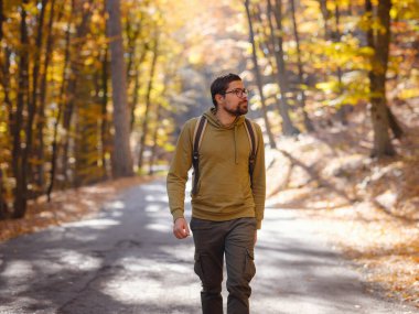 Genç yakışıklı adam sonbahar ormanında poz veriyor. Sırt çantalı genç hippi, ormanda duran gezgin, yürüyüş, orman, yolculuk, aktif sağlıklı yaşam tarzı, macera, tatil konsepti..