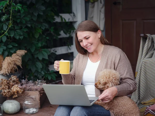 middle aged caucasian woman in brown cardigan sits on terrace of house with her beige poodle with laptop working outdoors in garden, home office , Education, modern lifestyle and leisure concept.