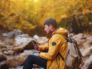 Macera ve aktif yaşam tarzı. Ormanda turist yürüyüşü. Sarı ceketli beyaz adam ormanda yürüyor. akıllı telefon, sosyal ağlar üzerinde navigasyon uygulaması kullanarak, resim çekerek