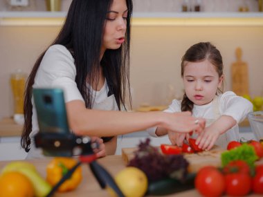 Happy mother and daughter enjoy prepare freshly salad together in kitchen. Healthy food at home. Healthy Lifestyle and Eating Concept. little influencer filming blog about healthy eating