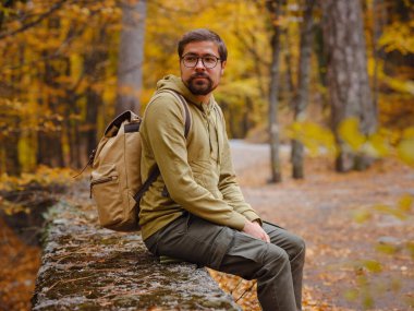 Genç yakışıklı adam sonbahar ormanında poz veriyor. Sırt çantalı genç hippi, ormanda duran gezgin, yürüyüş, orman, yolculuk, aktif sağlıklı yaşam tarzı, macera, tatil konsepti..