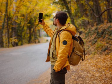 Sonbahar ormanında akıllı telefonlu genç yakışıklı bir adam. Sırt çantalı genç hippi, ormanda duran gezgin, yürüyüş, orman, yolculuk, aktif sağlıklı yaşam tarzı, macera, tatil konsepti..