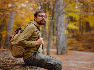 Genç yakışıklı adam sonbahar ormanında poz veriyor. Sırt çantalı genç hippi, ormanda duran gezgin, yürüyüş, orman, yolculuk, aktif sağlıklı yaşam tarzı, macera, tatil konsepti..