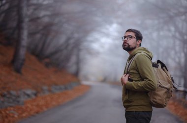 Genç yakışıklı adam sonbahar ormanlarında yolda poz veriyor. Sırt çantalı genç hippi, ormanda duran gezgin, yürüyüş, orman, yolculuk, aktif sağlıklı yaşam tarzı, macera, tatil konsepti..