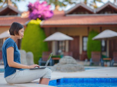 Young business woman working at computer by hotel pool. Young lady downshifter working at laptop and enjoys and relaxed environment, working day. Online freelance work on vacation.