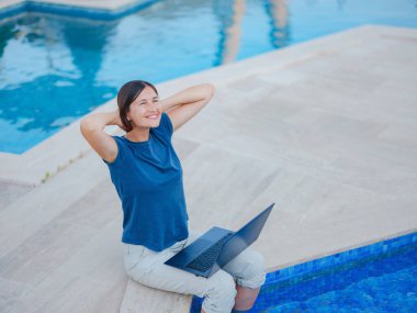 Young business woman working at computer by hotel pool. Young lady downshifter working at laptop and enjoys and relaxed environment, working day. Online freelance work on vacation.