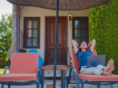 Young business woman working at computer by hotel pool. Young lady downshifter working at laptop and enjoys and relaxed environment, working day. Online freelance work on vacation.
