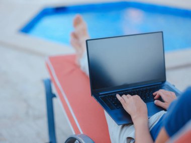 Young business woman working at computer by hotel pool. Young lady downshifter working at laptop and enjoys and relaxed environment, working day. Online freelance work on vacation.