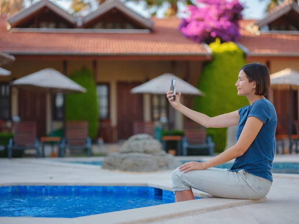 Young brunette woman using her phone while relaxing by the pool . Young lady downshifter working at phone enjoys and relaxed environment, working day. Online freelance work on vacation.