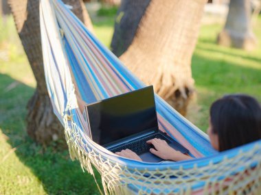 Young business woman working in a hammock. Young lady downshifter working at laptop and enjoys and relaxed environment, working day. Online freelance work on vacation.