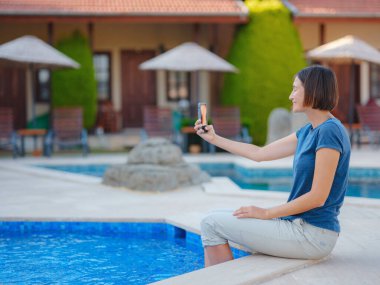 Young brunette woman using her phone while relaxing by the pool . Young lady downshifter working at phone enjoys and relaxed environment, working day. Online freelance work on vacation.