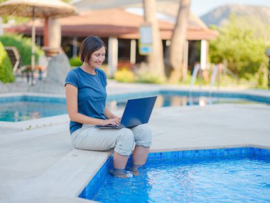 Young business woman working at computer by hotel pool. Young lady downshifter working at laptop and enjoys and relaxed environment, working day. Online freelance work on vacation.