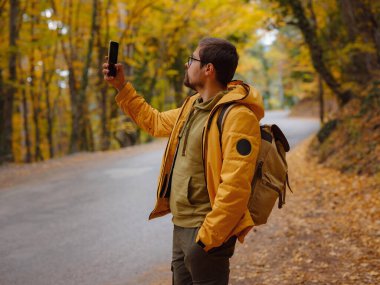 Sonbahar ormanında akıllı telefonlu genç yakışıklı bir adam. Sırt çantalı genç hippi, ormanda duran gezgin, yürüyüş, orman, yolculuk, aktif sağlıklı yaşam tarzı, macera, tatil konsepti..