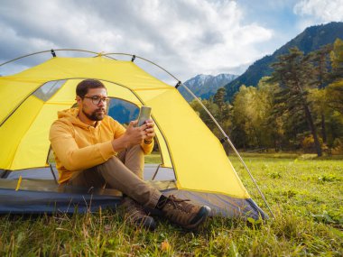 trip to Caucasus mountains, Arkhyz, Teberdinsky reserve. Man traveler relaxing in mountains in tent camping outdoor , uses app on smartphone. Travel adventure lifestyle concept hiking active vacations