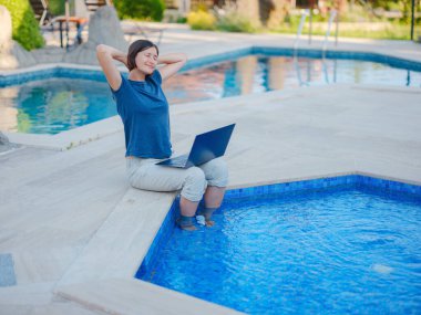Young business woman working at computer by hotel pool. Young lady downshifter working at laptop and enjoys and relaxed environment, working day. Online freelance work on vacation.