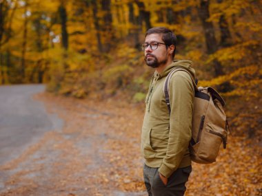 Genç yakışıklı adam sonbahar ormanlarında yolda poz veriyor. Sırt çantalı genç hippi, ormanda duran gezgin, yürüyüş, orman, yolculuk, aktif sağlıklı yaşam tarzı, macera, tatil konsepti..