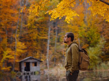 Young handsome man posing in autumn forest near lake. young hipster guy with backpack , traveller standing in woods, Hiking, Forest, Journey, active healthy lifestyle, adventure, vacation concept.