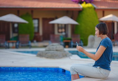 Young brunette woman using her phone while relaxing by the pool . Young lady downshifter working at phone enjoys and relaxed environment, working day. Online freelance work on vacation.