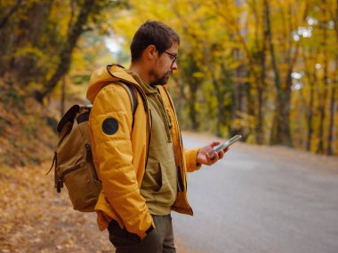 Young handsome man in autumn forest with smartphone. young hipster guy with backpack , traveller standing in woods, Hiking, Forest, Journey, active healthy lifestyle, adventure, vacation concept.
