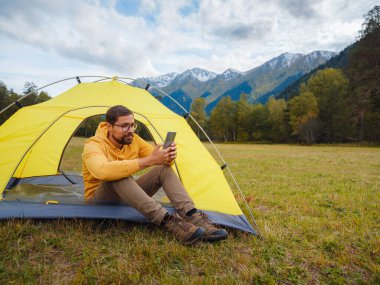 trip to Caucasus mountains, Arkhyz, Teberdinsky reserve. Man traveler relaxing in mountains in tent camping outdoor , uses app on smartphone. Travel adventure lifestyle concept hiking active vacations