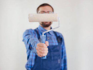 Mavi takım elbiseli genç adam beyaz duvar arkasında izole edilmiş boya silindiri tutuyor. Yenileme dairesi için aksesuarlar. Ev konseptini onar