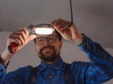 Elektrikçi iş başında. Elektrik kablolarının tamiri ve tavan lambalarının değiştirilmesi için servis. İnşaatçı tavana küçük bir lamba yerleştiriyor. Kiralık centilmen ev işlerine yardım eder.