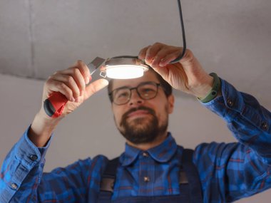 Elektrikçi iş başında. Elektrik kablolarının tamiri ve tavan lambalarının değiştirilmesi için servis. İnşaatçı tavana küçük bir lamba yerleştiriyor. Kiralık centilmen ev işlerine yardım eder.