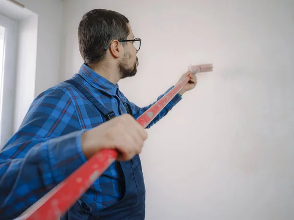 young man in blue work suit hold paint roller, over white wall room background. Instruments accessories for renovation apartment room. Repair home concept