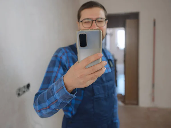 young man in blue work suit doing Repair apartment. Home renovation concept. Guy with smartphone texting and tapping on cellphone, searching for solution on Internet, ordering goods for repairs