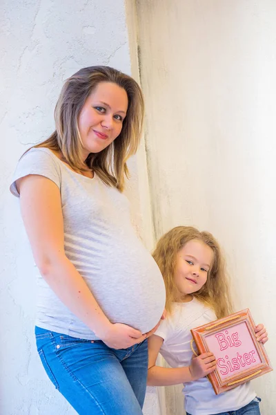 Beautiful pregnant woman with daughter standing near a wall at home — Stock Photo, Image