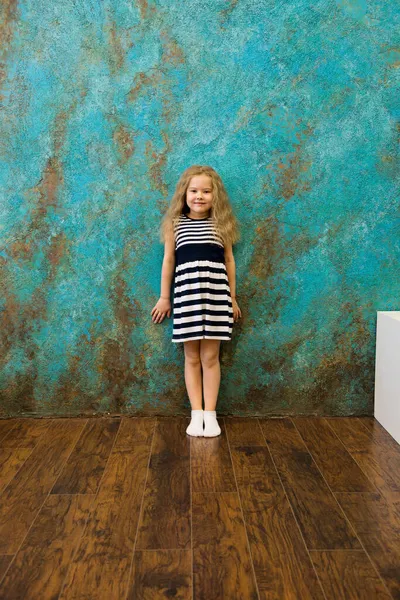 Menina bonito em roupas listradas de pé perto da parede — Fotografia de Stock