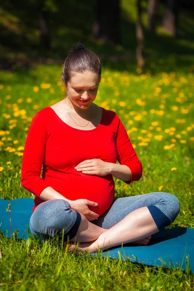 Pregnancy yoga exercise - pregnant woman doing asana Sukhasana easy yoga pose holding her abdomen outdoors on grass lawn with dandelions in summer