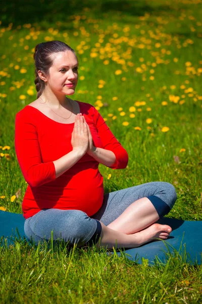 Pregnancy yoga exercise - pregnant woman doing asana Sukhasana easy yoga pose with namaste salutation outdoors on grass lawn with dandelions in summer