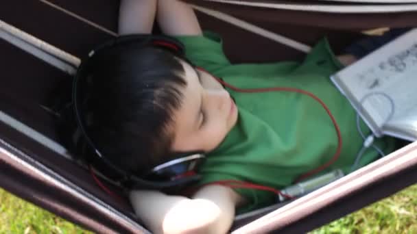 Мальчик читает и слушает музыку в гамаке — стоковое видео