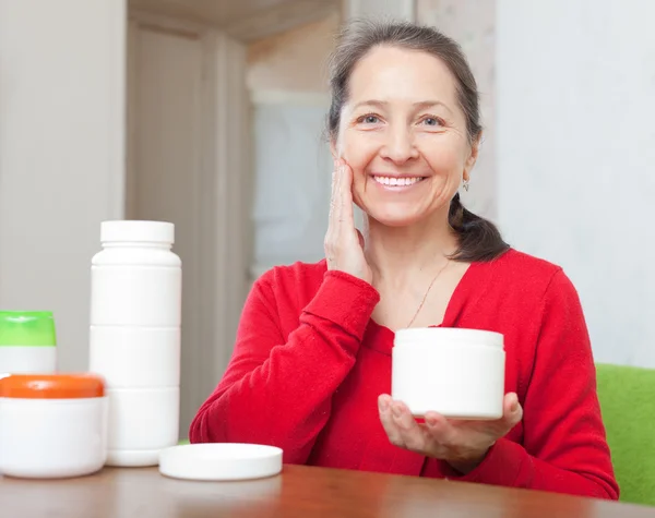 Smiling mature woman puts cream on face — Stock Photo, Image