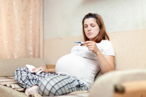 Donna incinta seduta sul divano con termometro — Foto Stock