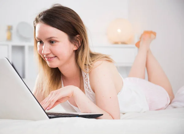 Söt flicka i sovkläder som ligger med laptop på sängen och arbetar — Stockfoto