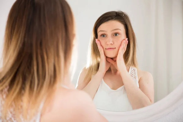 Młoda kobieta za pomocą lustra i dotykając twarzy w sypialni — Zdjęcie stockowe